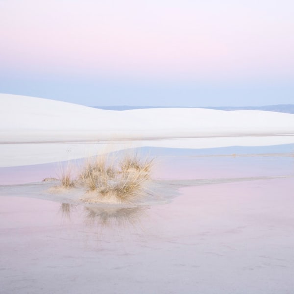 A landscape of white sand dunes with the pink and blue colors of a sunset reflecting on water.