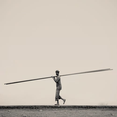 Man walking in the center of an empty landscape carrying a long pieces of wood on his shoulder.
